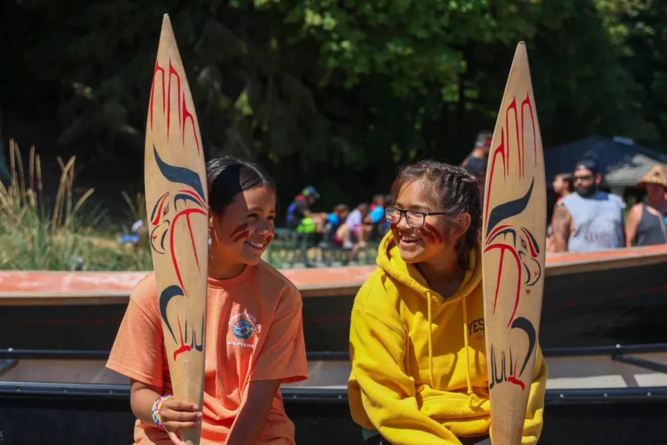 Two young girls are smiling with red face paint on. One is wearing a orange shirt and the other girl is wearing glasses and a yellow hoodie.